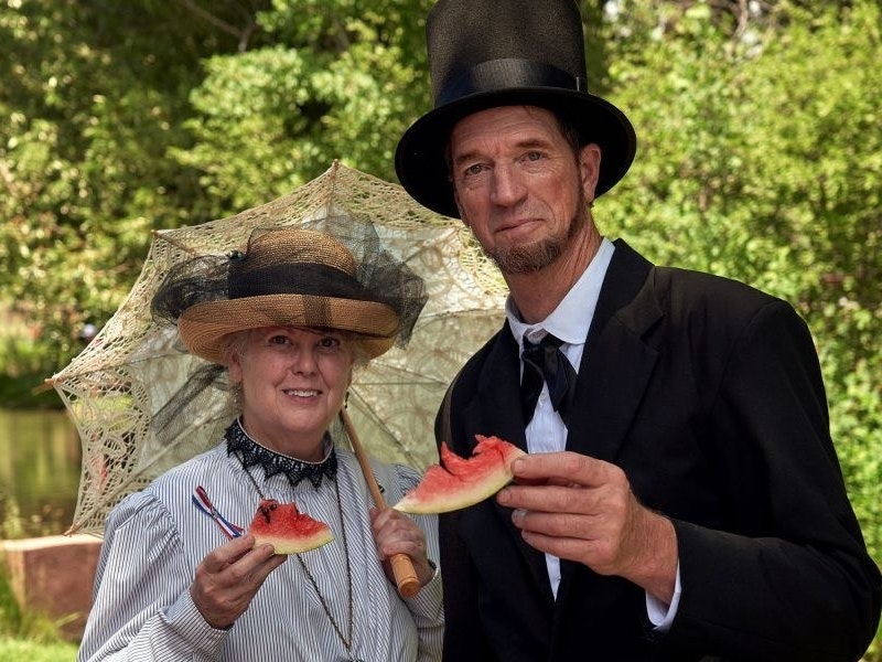 Actor portraying Abraham Lincoln and a lady dressed in vintage period dress and hat with parasol with green trees in the background.